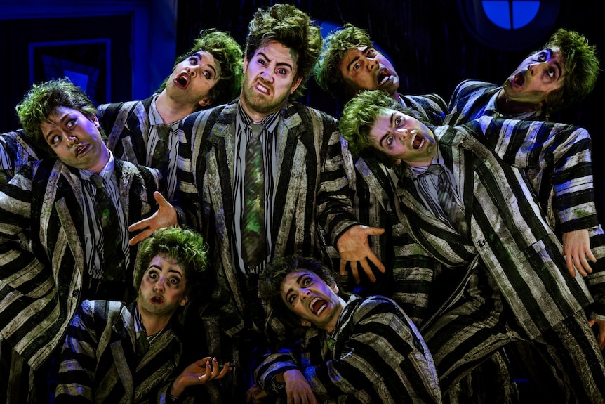 On stage, Eddie Perfect, in a prison-striped suit, poses, grimacing, an ensemble of similarly dressed actors around him.
