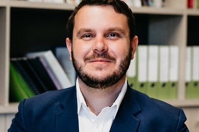 A midrange head shot of a man wearing a navy jacket and white shirt, with brown hair and a trimmed beard.