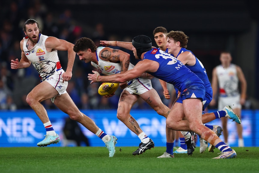 A footballer runs with the ball under his right arm and is about to be tackled