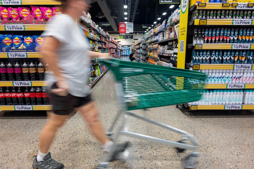 A person wheels their trolley past the end of a supermarket aisle. They are blurred from the motion and are not identifiable.