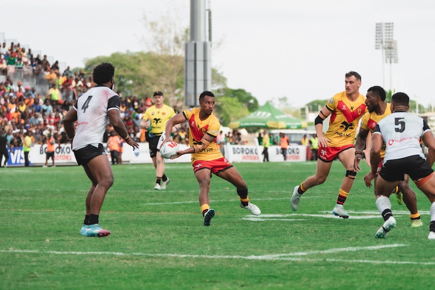 A PNG man in yellow and red rugby league uniform goes to pass a ball on a rugby field facing men in white