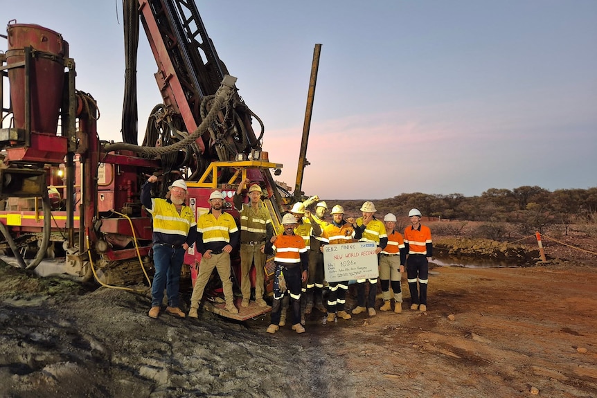 Men in hard hats and high-vis stand in front of a drill rig in an outback area.
