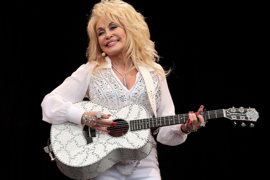 Dolly Parton smiling, playing guitar in a sparkling suit.
