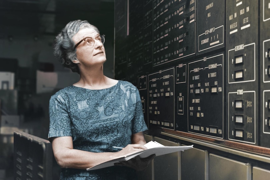 A woman holds a sheaf of papers and pencil while looking up at a wall of labelled computer equipment.