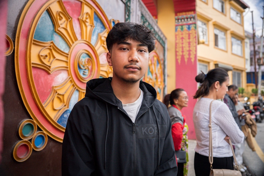 A young man stands in front of an ornate gate. He's wearing a black hooded jacket.