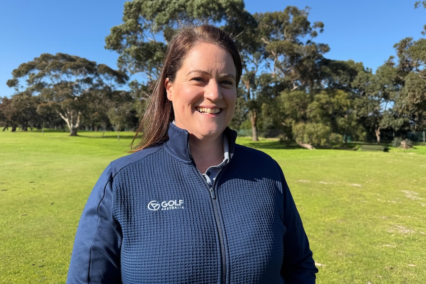 Tamara Mason smiles at the camera. She has brown hair and a navy jacket. She stands outside on a green golf course.