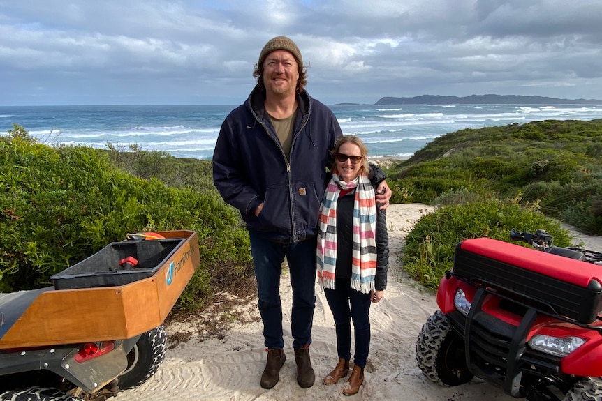 Longley stands towering over Shea as they stand in sand dunes with a beach in the background.