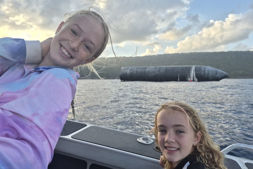Two blonde girls smile from the side of a boat, with the SpaceX rocket behind them. 