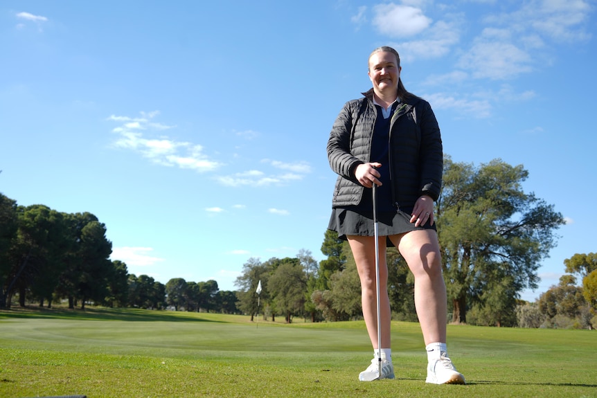 Melissa Miegel stands facing the camera holding a golf club in front of her. She is on a golf course, wearing a black jacket.