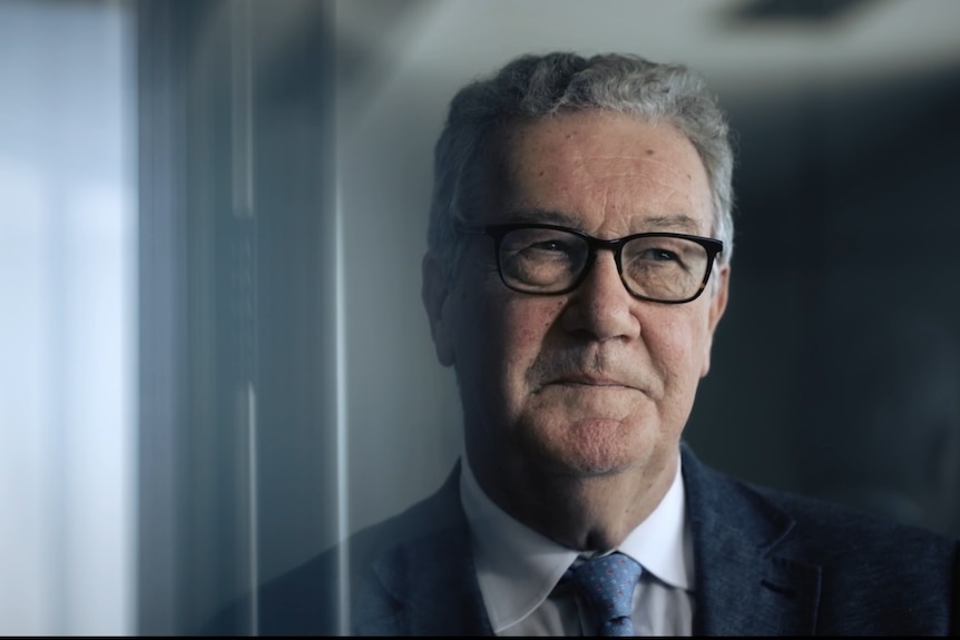A medium close-up shot of an older man with grey hair, glasses and a wry expression.
