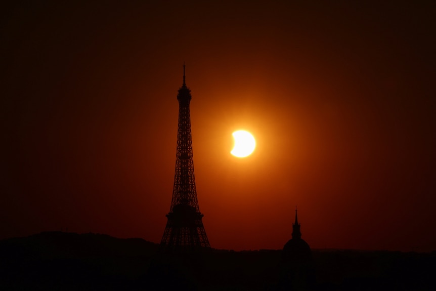 The partially eclipsed Sun sits behind the Eiffel Tower. The sky is a dark red.