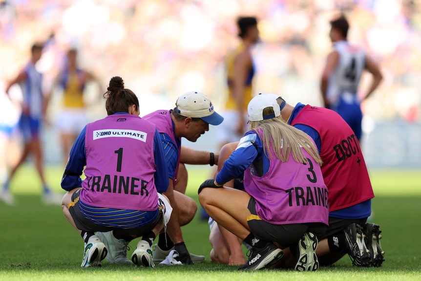 Trainers and a club doctor surround an AFL player on the ground, who is obscured.