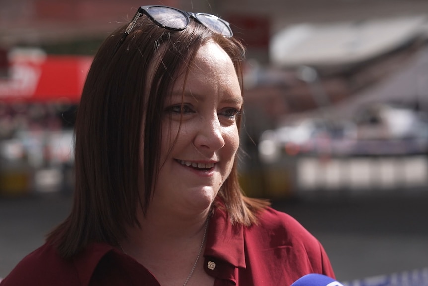 A woman with a bob wearing a shirt and glasses on her head talks to the media in frotn of a service station.