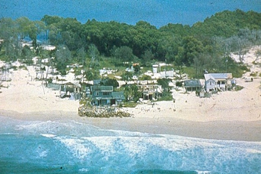 An old aerial photo showing damaged houses, surrounded by sand and rock with beach in front and a creek behind.