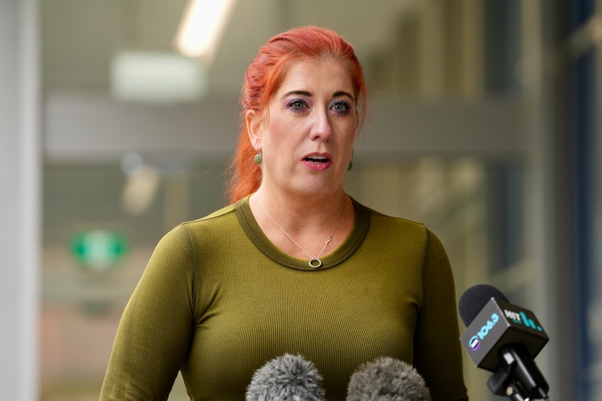 A woman in a green shirt speaks with a serious expression on her face.