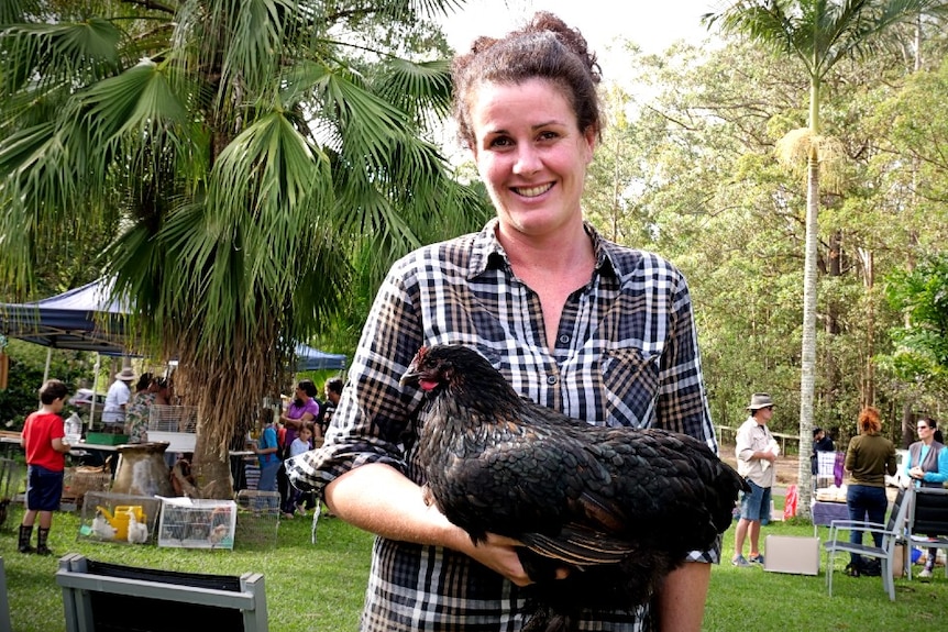 A smiling, dark-haired woman in a checked shirt holds a dark hen while standing in an outdoor market.