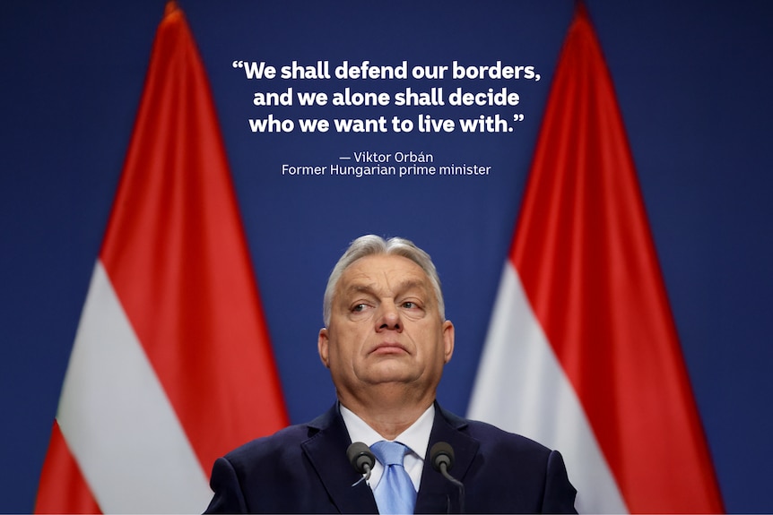 A white man with grey hair wears a black suit and light blue tie, with two large Hungarian flags behind.