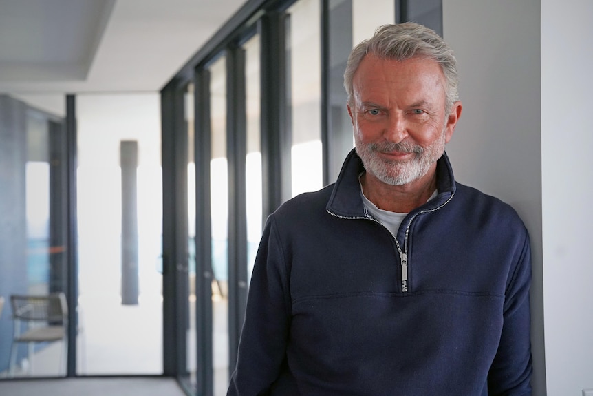 Sam Neill leans against glass sliding doors in an apartment.