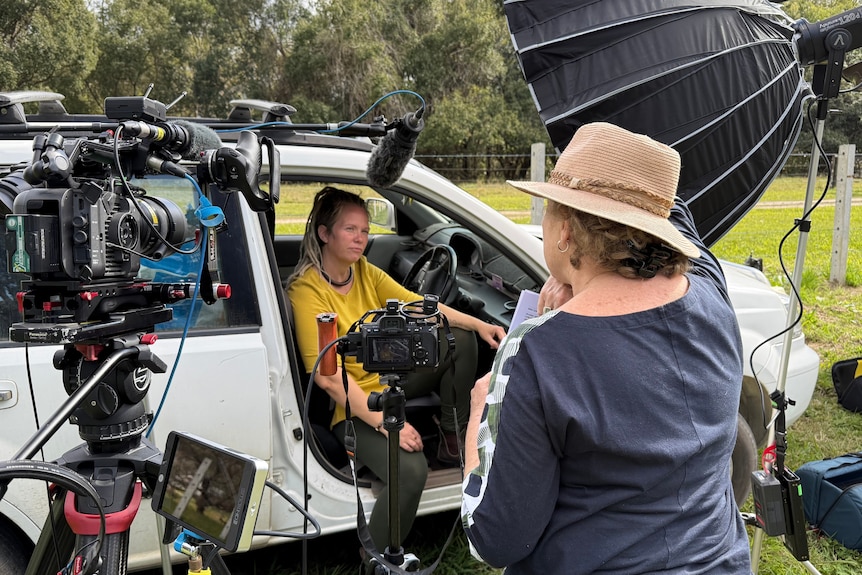 A woman in a yellow top sits in a car with the door open. TV cameras and a woman are in front of the door.
