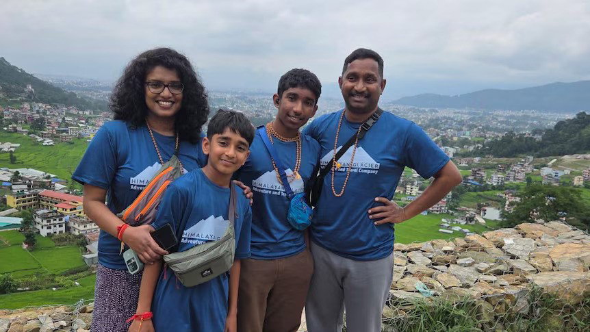 A family of four people, all wearing blue shirts and cross body bags standing at a site, with greenery behind them.