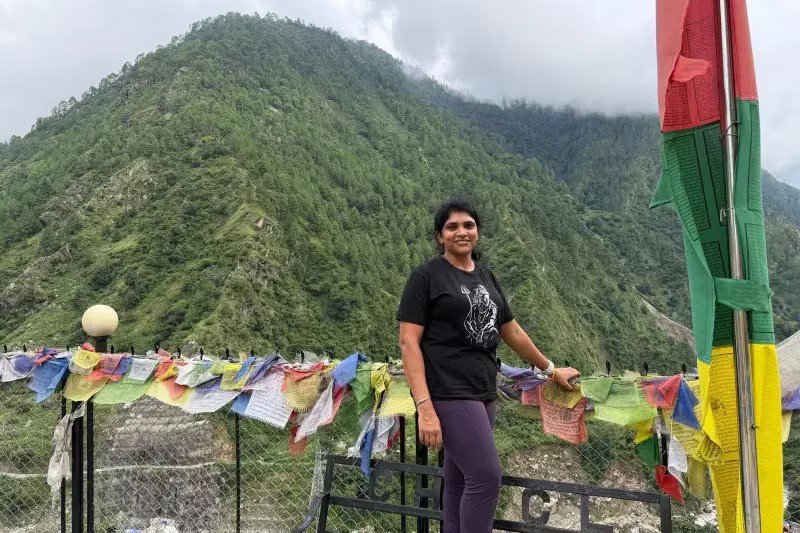 A woman standing in tshirt and leggings at a gate with a mountain top behind her.