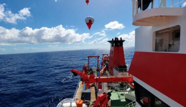 View from a research vessel coloured red and white, that is sailing on open sea, on two helikites that rise into the blue sky.