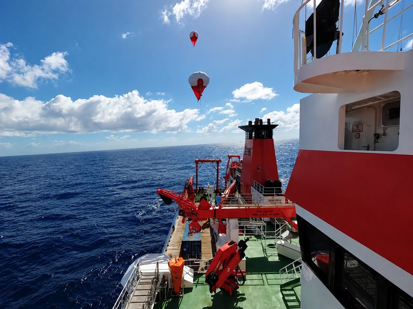 View from a research vessel coloured red and white, that is sailing on open sea, on two helikites that rise into the blue sky.