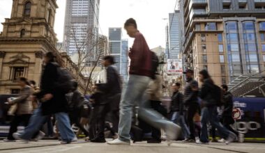 Blurred pedestrians crossing a busy Melbourne city street, with trams, historic buildings and modern skyscrapers in the background.