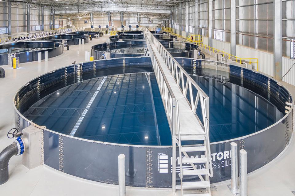 Interior view of a large industrial aquaculture or water treatment facility containing multiple circular tanks filled with water. A central metal walkway extends across the nearest tank, with additional tanks, pipes, railings, and overhead structural beams visible throughout the spacious warehouse-like building.
