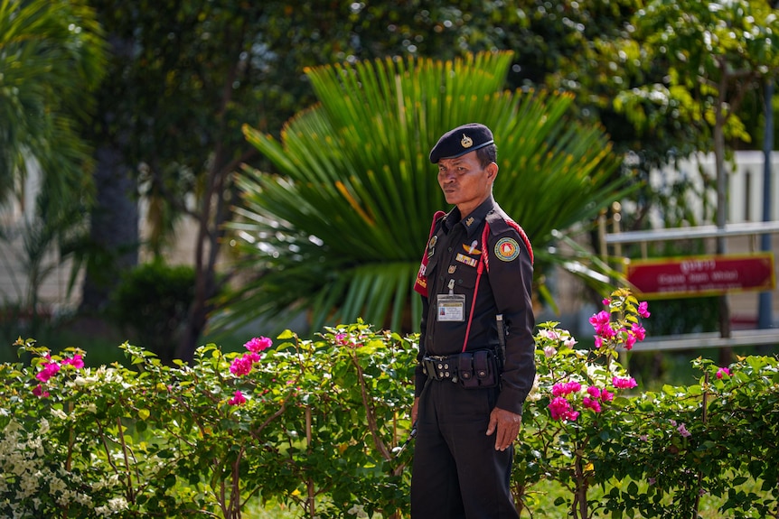 A man in a dark, military-style uniform stands in front of a large garden.