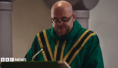 Father Luke wears a green chasuble with yellow brocade. He has a shaved head and dark round glasses.