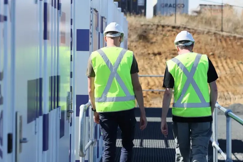 Two men in hard hats and fluro vests walk away from the camera, alongside a shipping-container-like wall.