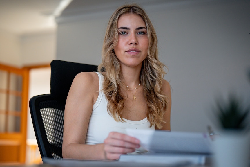 A young woman with blonde curly hair sits at a desk with an open textbook and a computer
