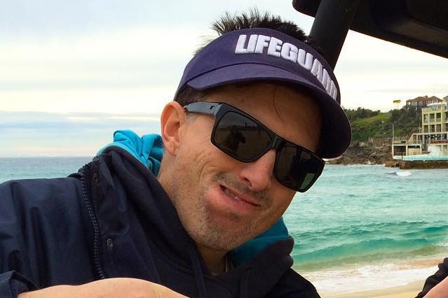 A man wearing black sunglasses, a towel poncho and 'Lifeguard' visor pointing to one side with his tongue pointing out.
