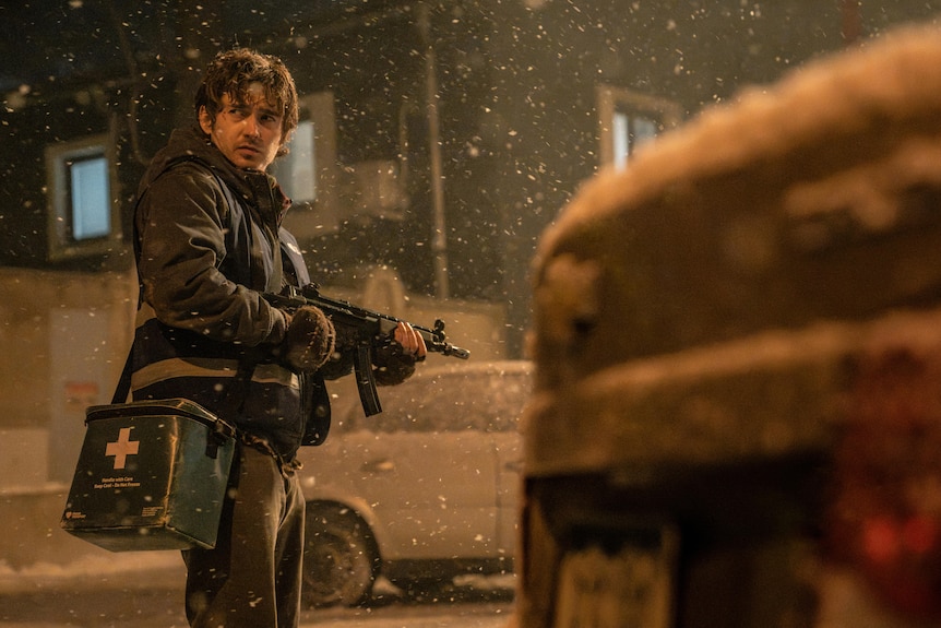 A man with a gun stands near some cars in the snow.