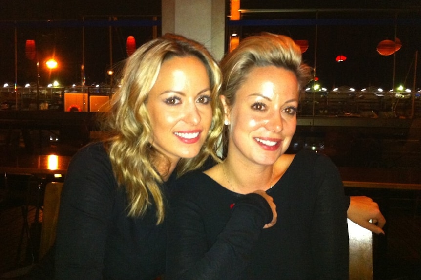 Two women with blonde hair and black shirts smile at the camera. They are twins.