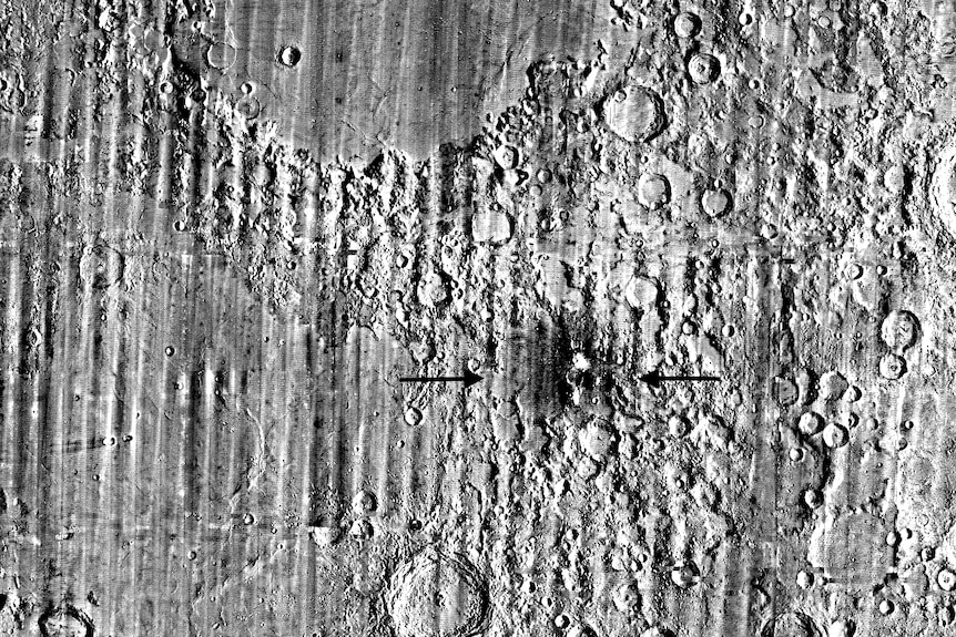 A greyscale image of dozens of Moon craters, with a particular hole marked by two black arrows.