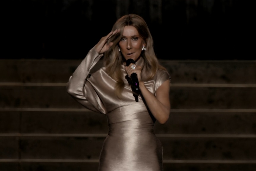 Celine holds a mic and raises her hand to her face. She wears a bronze, shiny dress.