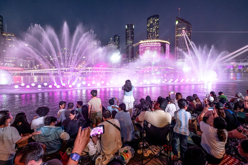 A water and light show on a river at night being watched by people on the river shore.