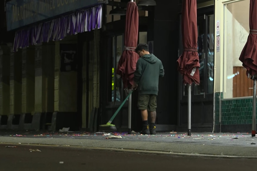 A man sweeps a footpath littered with rubbish outside a pub at night.