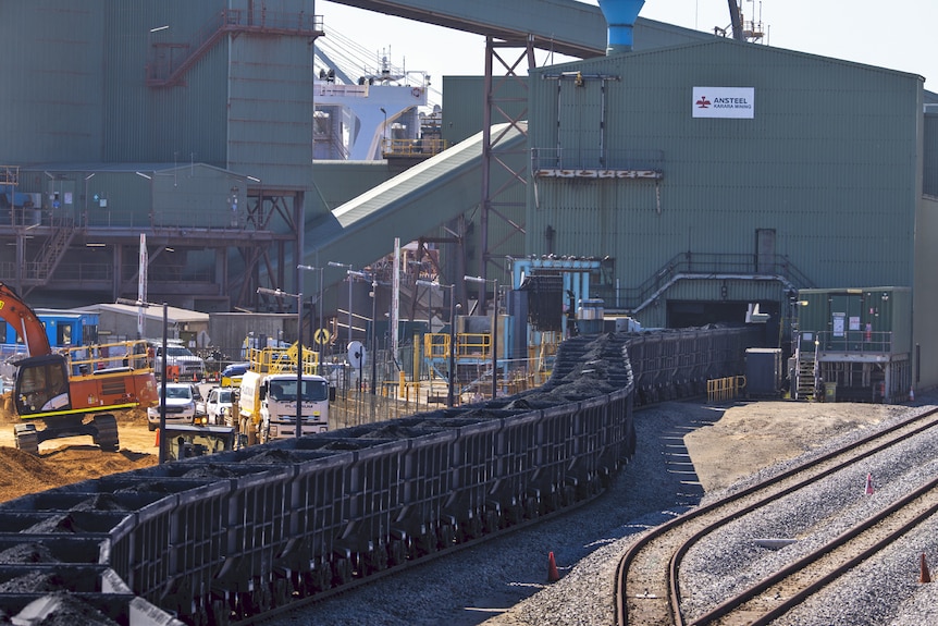 A wide shot of a long train carriage loading ore into the large green and grey Karara shed ready for export.