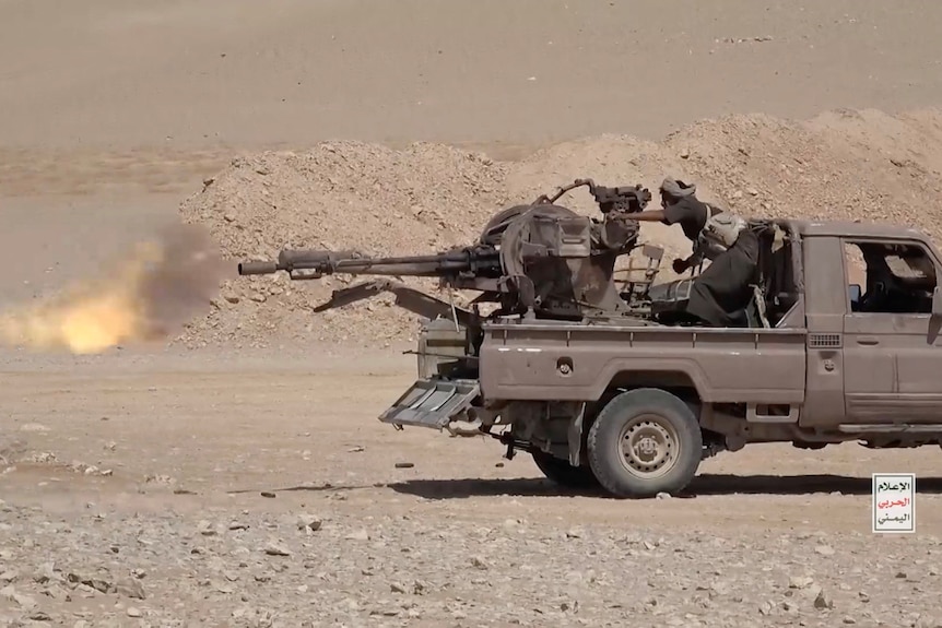 A ute with a machine gun mounted on the back tray firing in a desert.