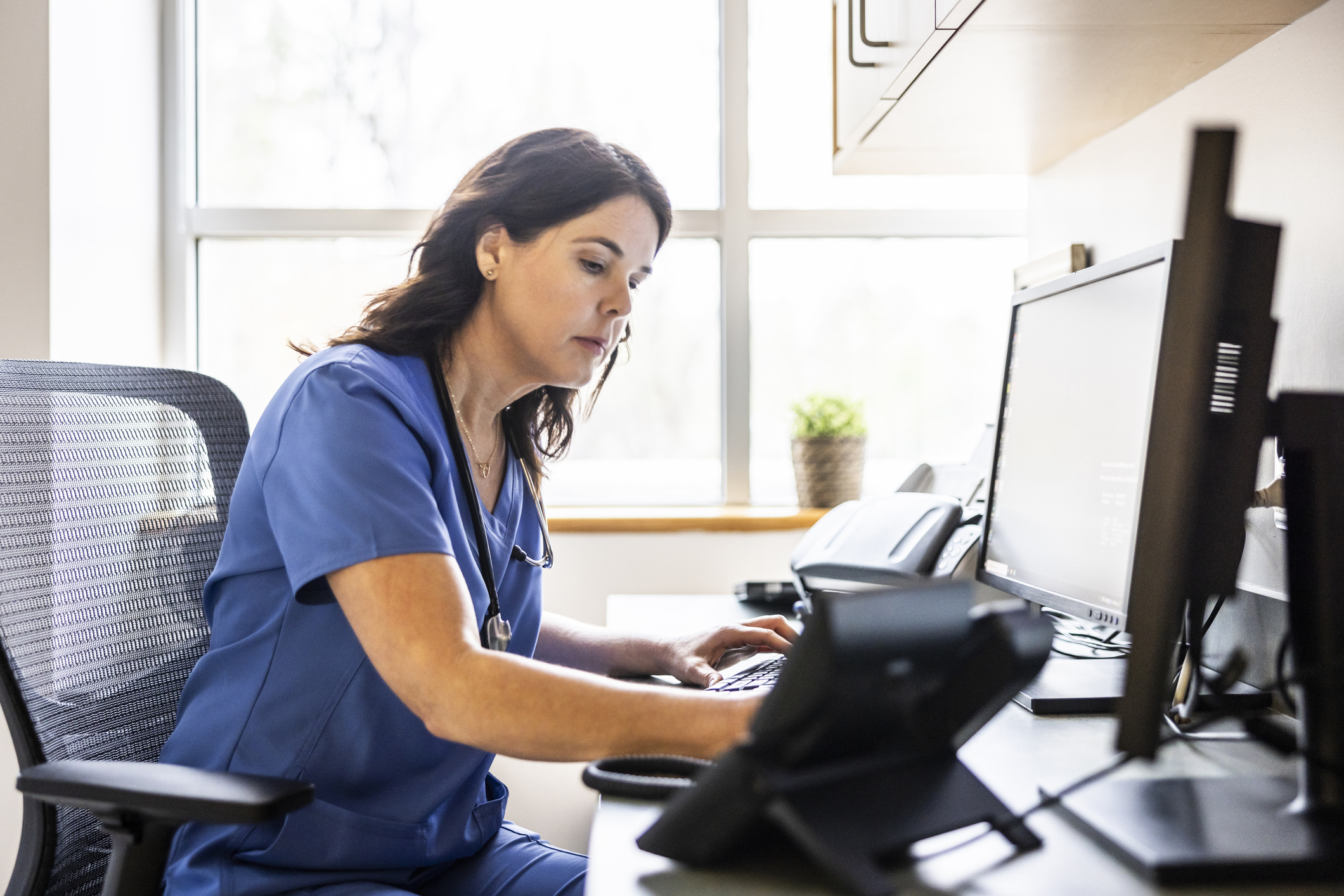 A person in blue scrubs with long brown hair checking messages on a desktop computer.