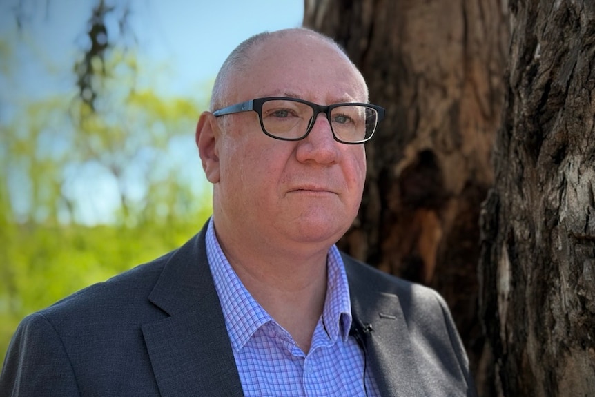A middle-aged man in glasses and a dark suit stands next to a large tree.