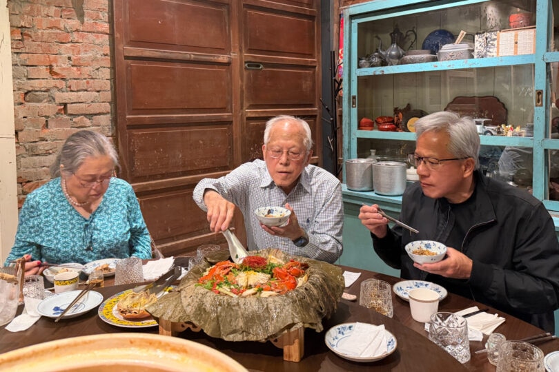 Jensen Huang in a black jacket eats from a bowl with chopsticks at a table with an elderly man and woman.
