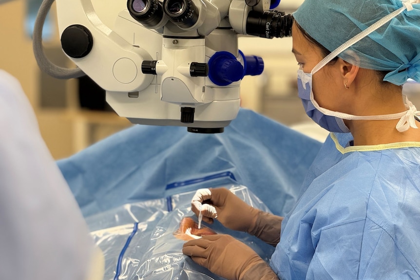 A female surgeon in scrubs stands beside a patient on an operating table, with only their right eye exposed.
