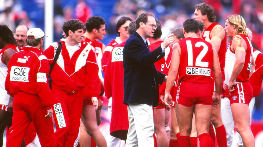 A Sydney Swans AFL coach stands on the SCG gesturing as he talks to a group of players during a game.