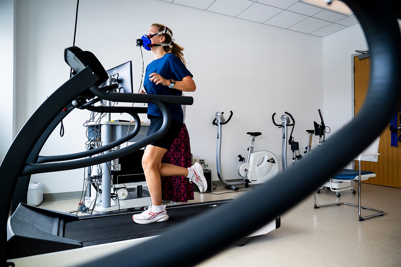 A student runs on a treadmill wearing a respiratory mask connected to monitoring equipment, shot through the frame of another treadmill in the foreground.