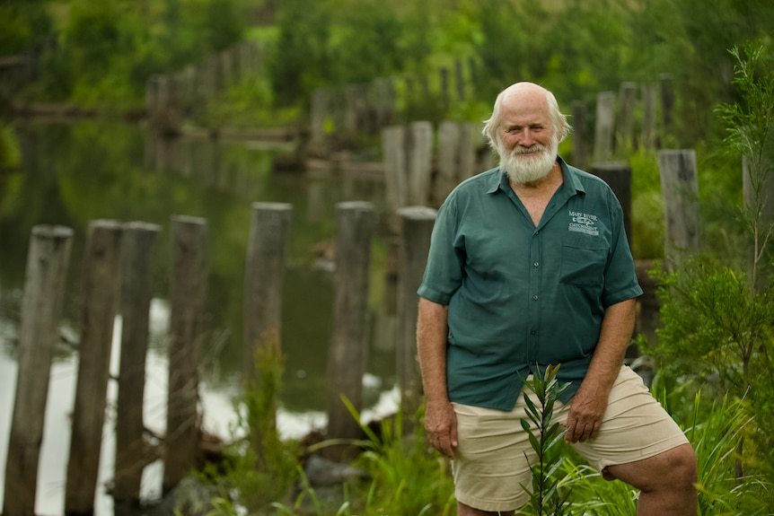 A man with a white beard, dark green short sleeved shirt and khaki pants stands in front of rows of timber in a creek bed.