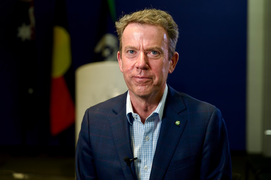 A middle aged man wearing a dark blue suit standing in front of Australian and Aboriginal flags
